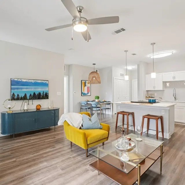 A modern and bright living area featuring a yellow accent chair, coffee table, and a sleek TV stand with a landscape painting above it. The kitchen is open and equipped with white cabinetry and stainless steel appliances.