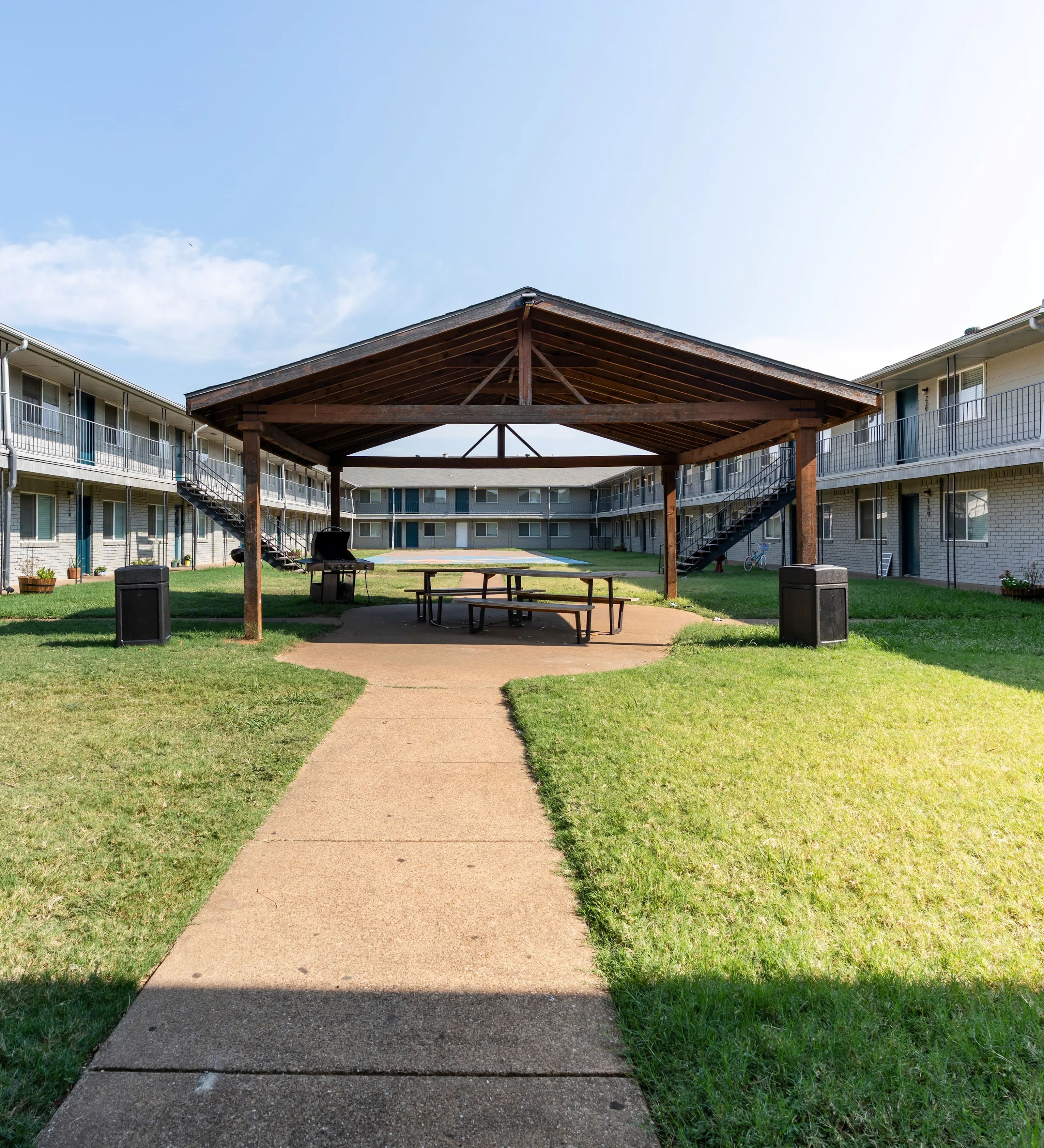 Courtyard Pavilion in Apartment Complex A paved pathway leads through a grassy area with a wooden pavilion at the center. The pavilion features a table underneath it, and there are two speakers on either side. Surrounding the pavilion are two-story buildings with balconies.
