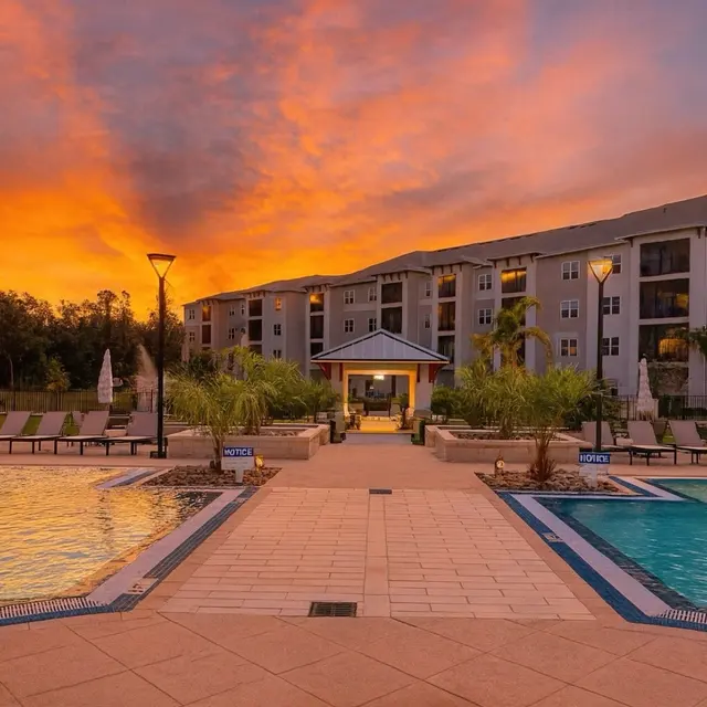 A tranquil pool area at sunset with two swimming pools surrounded by lounge chairs and palm trees. In the background, there is a multi-story building under a vibrant orange and purple sky.