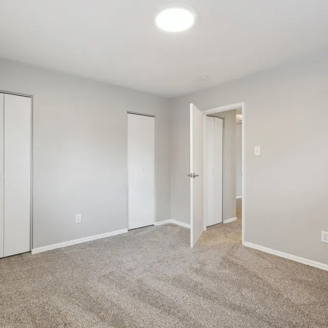 A spacious empty room with beige carpet, light gray walls, and a ceiling light. There are two closet doors on one wall and a partially opened door leading to another room.