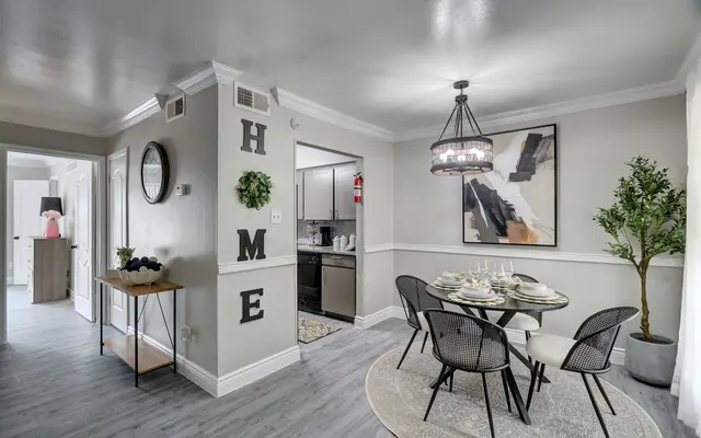 A modern dining area featuring a round table with place settings, surrounded by stylish black chairs. The walls are decorated with a wall art piece and the word 'HOME'. A large potted plant adds greenery to the space. The area is well-lit and has a contemporary feel.