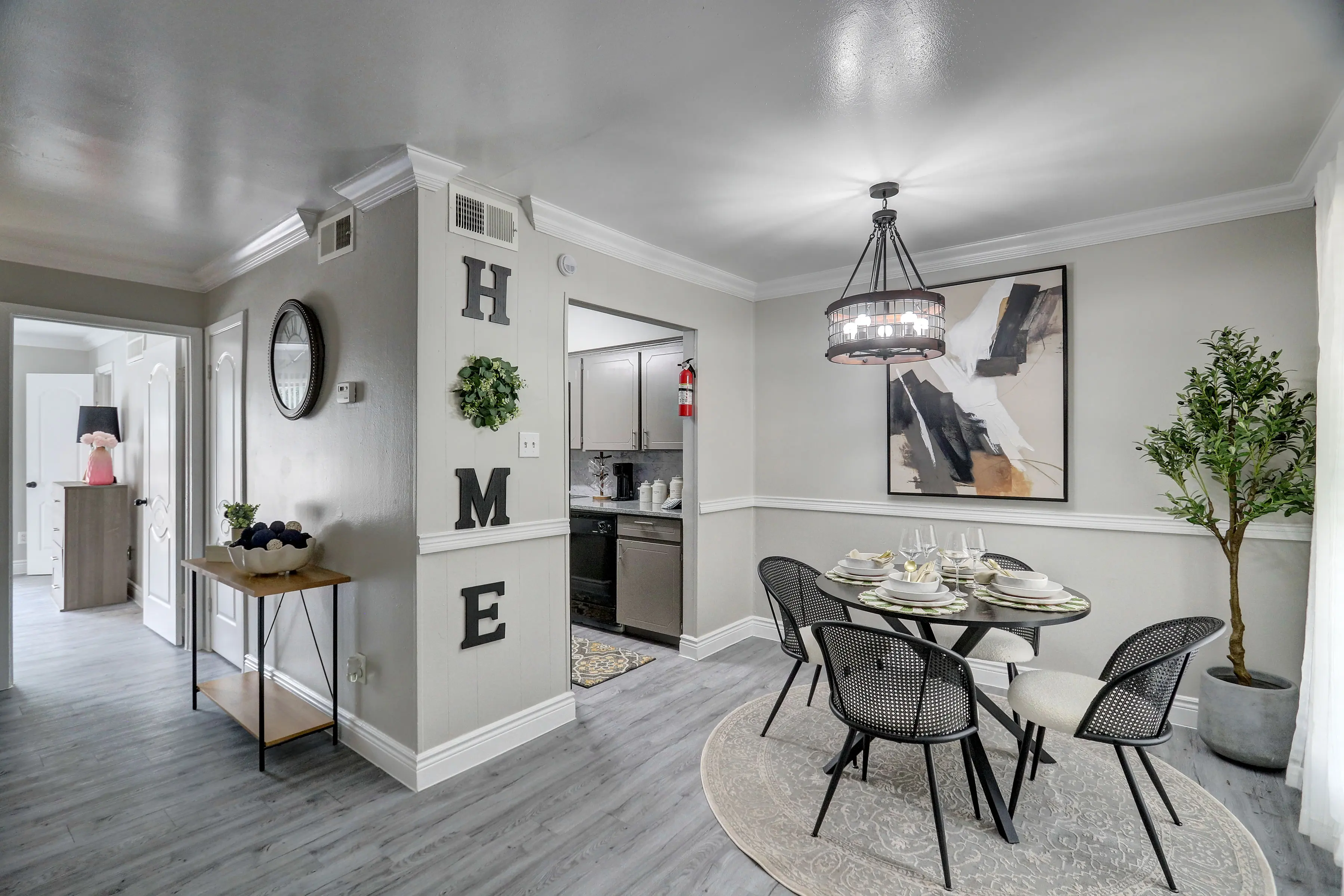 A modern dining area featuring a round table with place settings, surrounded by stylish black chairs. The walls are decorated with a wall art piece and the word 'HOME'. A large potted plant adds greenery to the space. The area is well-lit and has a contemporary feel.