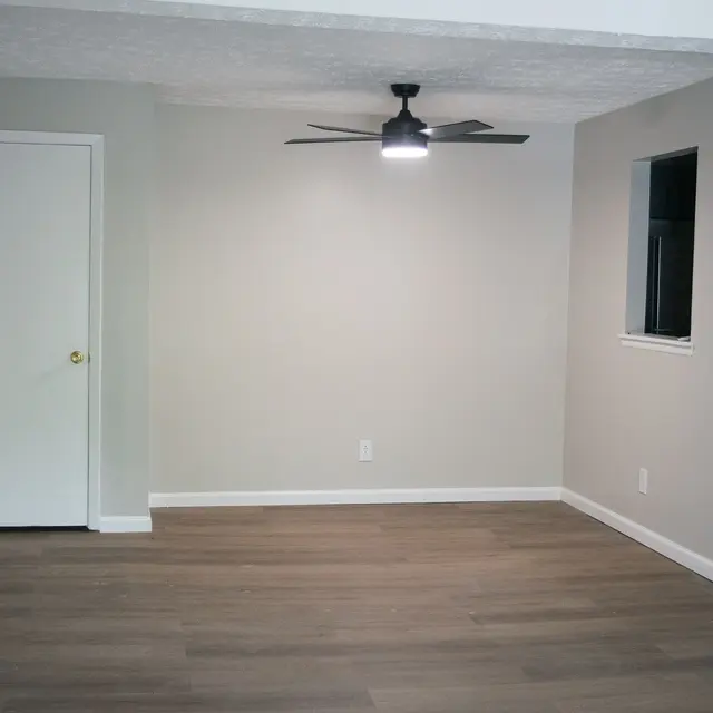 A spacious, empty room with light gray walls, wooden flooring, a ceiling fan, and a window, featuring a door on the left.