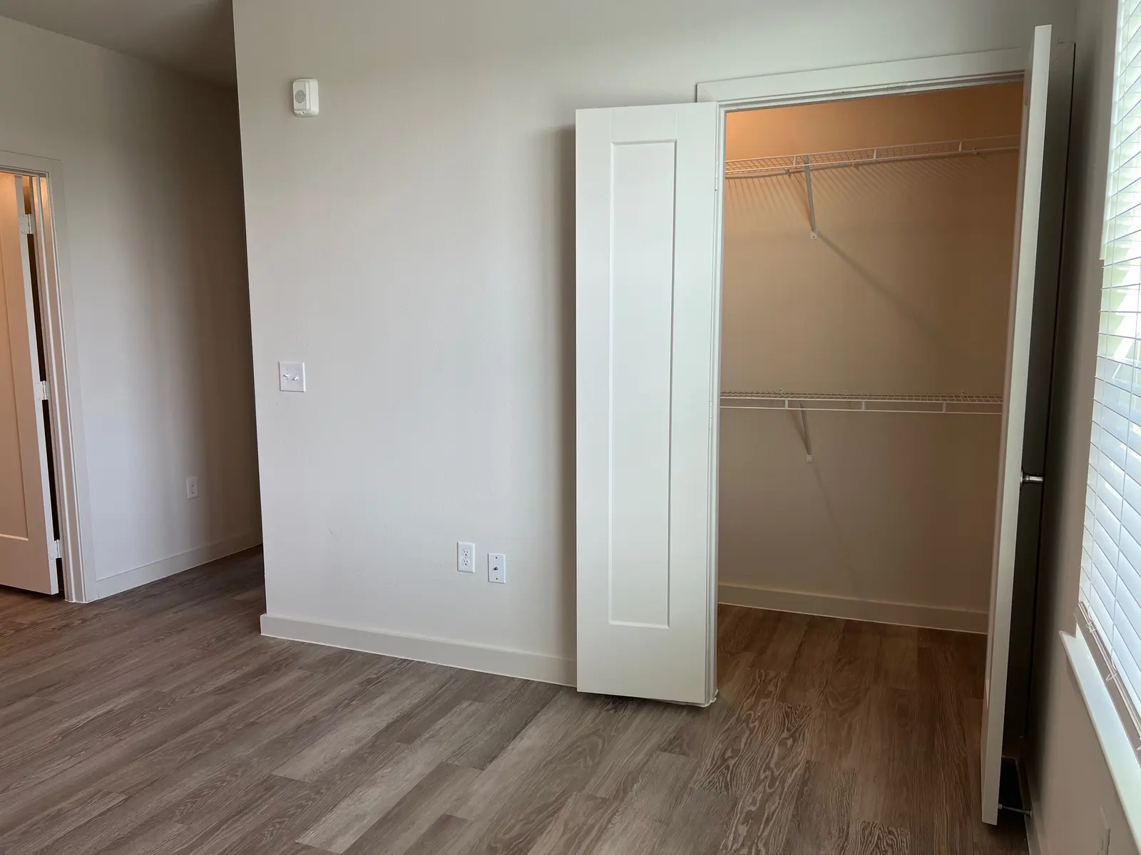 A spacious, empty room featuring light-colored walls and flooring. A closet with bi-fold doors is open, showcasing wire shelving inside. There is a window with blinds on one side, allowing natural light to enter the room.