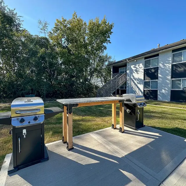 Outdoor Grilling Area An outdoor grilling area with two grills and a prep table on a concrete slab, surrounded by grass and trees, with an apartment building in the background.