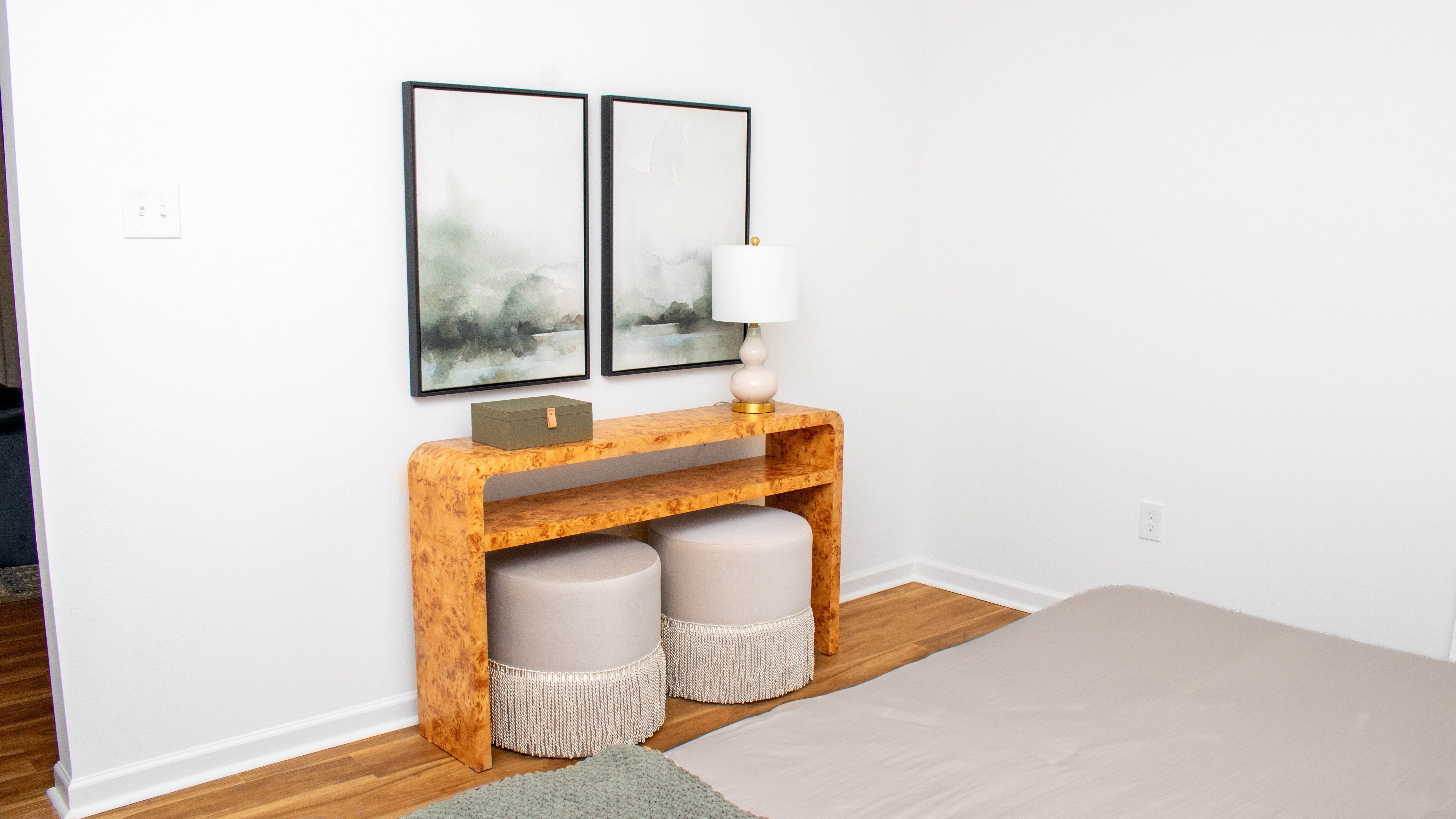 A minimalist bedroom scene featuring a light-colored bed and decor. A wooden console table with a lamp and two round stools with fringes are placed against a white wall. Above the table are two framed artworks depicting abstract landscapes.