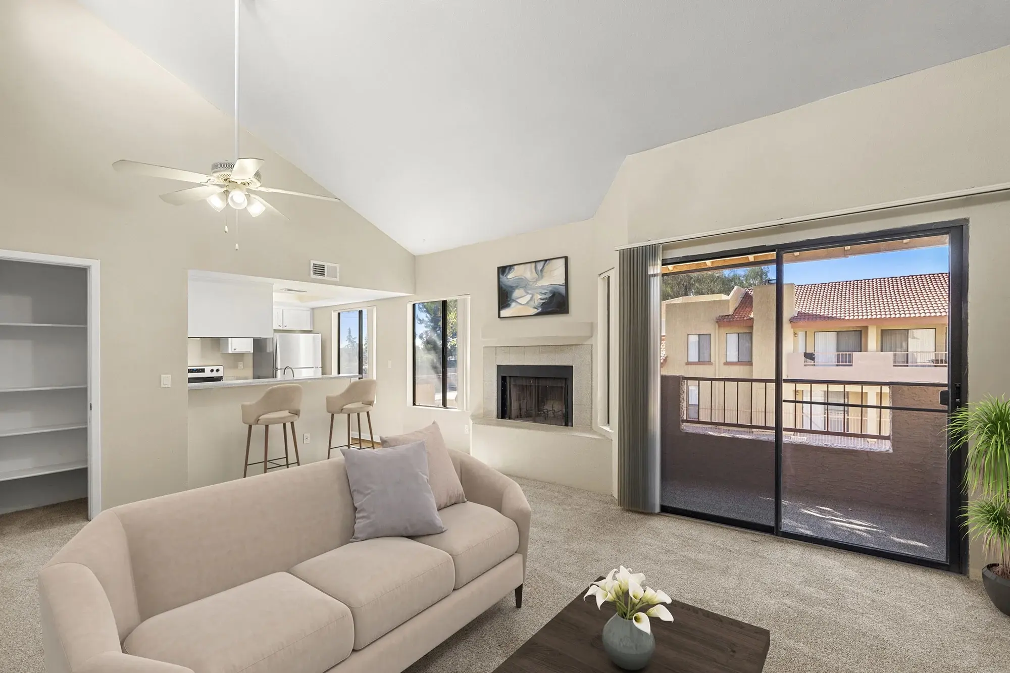 A spacious and bright living room featuring a beige couch, a wooden coffee table with a plant, and a balcony door. In the background, there is a kitchen area and a fireplace.