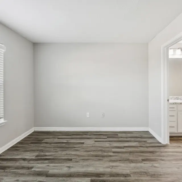 A light, empty room with gray walls, a window with blinds, and a doorway leading to a bathroom area. Wooden floor.