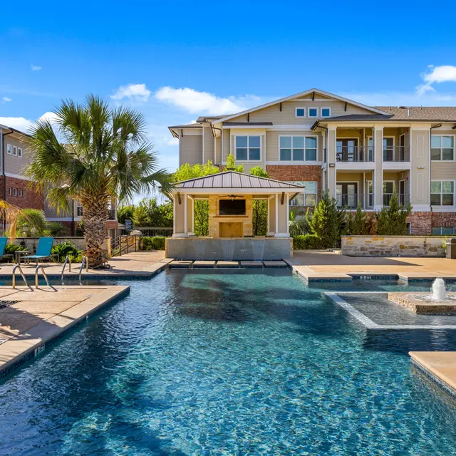 A luxury apartment complex with a swimming pool, palm trees, and outdoor lounge areas under a clear blue sky.