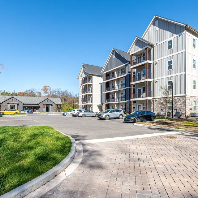 A modern multi-story apartment complex with balconies and a parking area. A small building in the background and a clear blue sky above.
