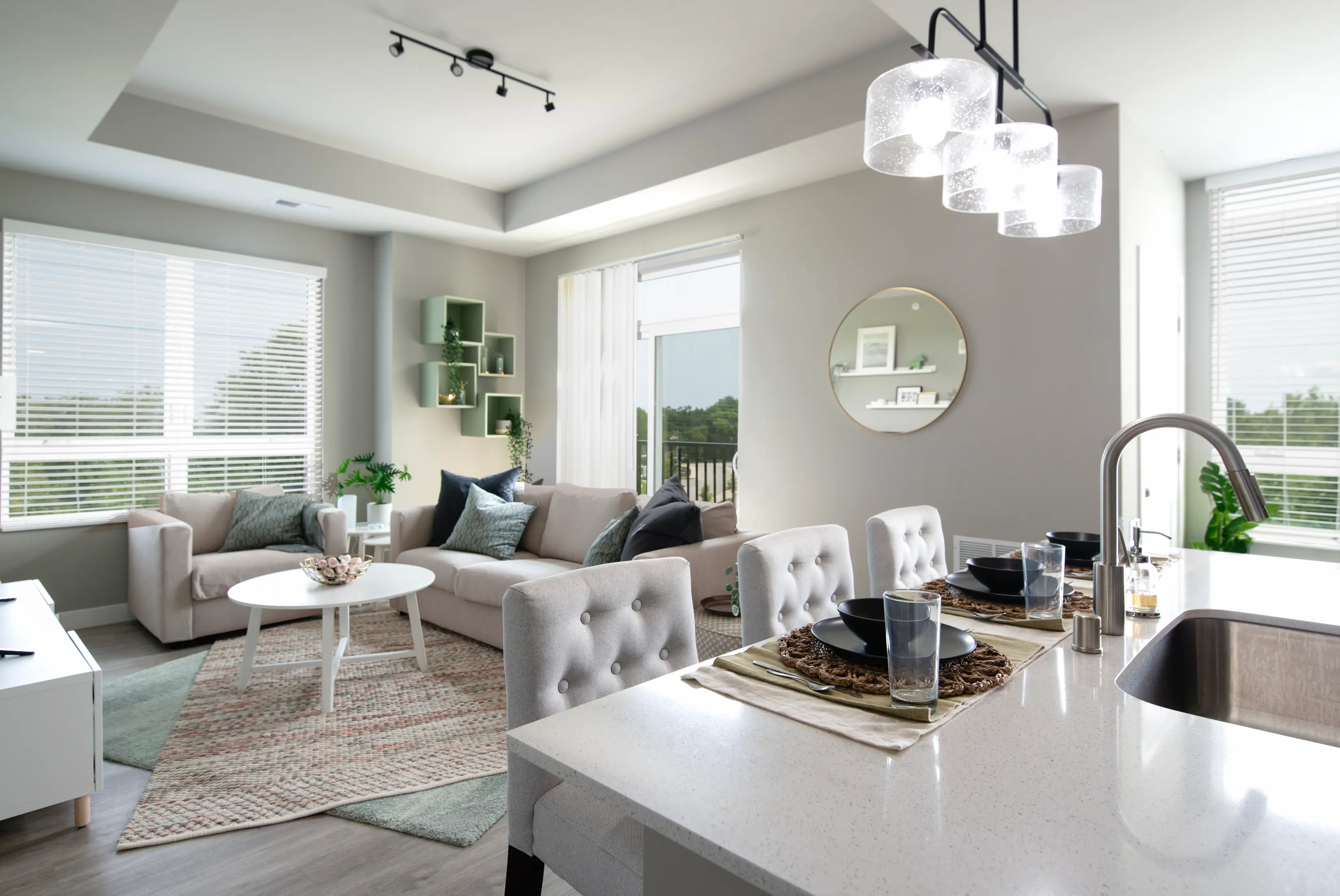 A modern and stylish living room with a comfortable beige sofa, round white coffee table, and green decor items. A dining area is visible with a sleek white table set with plates and glasses. The room has large windows with blinds letting in natural light, and a neutral color palette is used throughout.