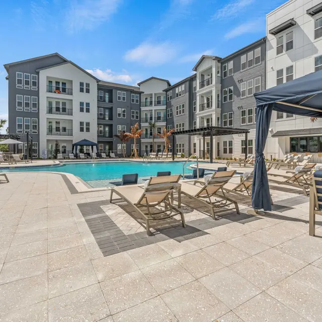 Modern Apartment Pool Area A modern apartment complex featuring a large swimming pool surrounded by lounge chairs and cabanas. The building has a sleek design with gray and white façades and large windows, set under a clear blue sky.