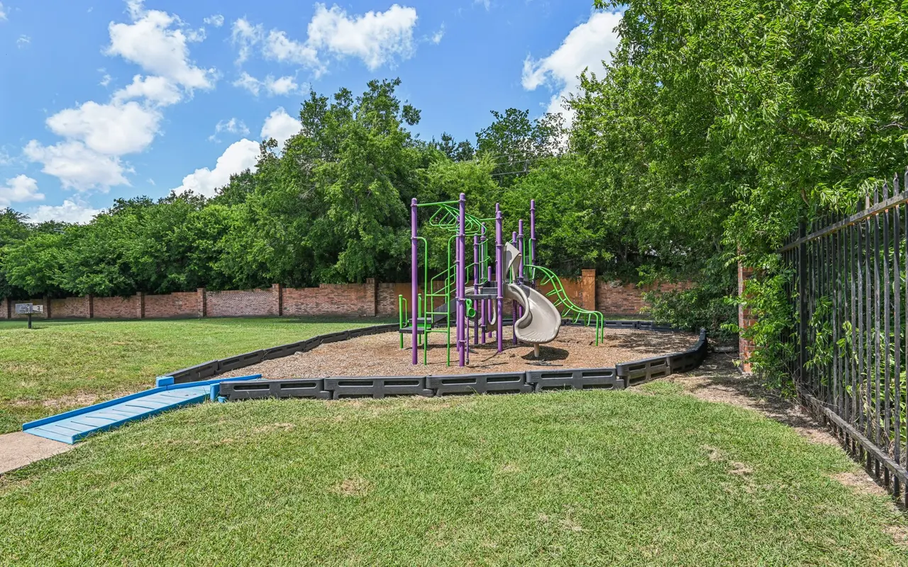 A playground featuring a purple slide and climbing structures surrounded by green grass and trees under a blue sky with fluffy clouds.
