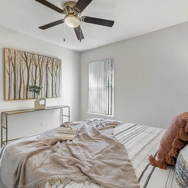 A cozy bedroom featuring a large bed with neutral-toned bedding and a warm throw, complemented by a decorative painting of trees on the wall. A console table is placed beside the bed, and there is a window with blinds allowing for natural light to enter the room.