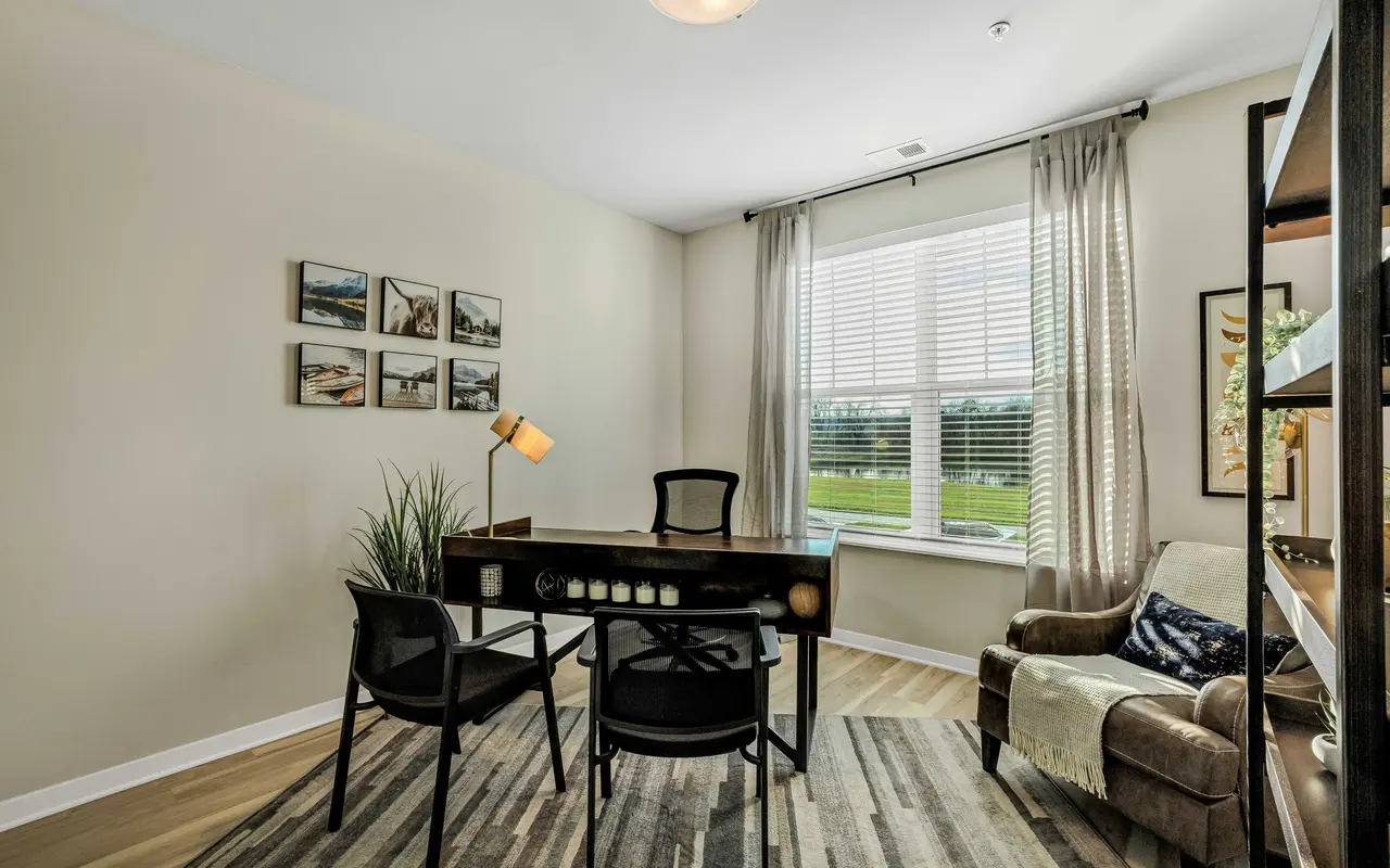 A modern home office featuring a desk with a lamp, two black chairs, a window with natural light, and decorative wall art.