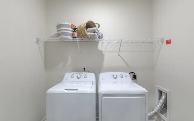 A clean and organized laundry room featuring a white washing machine and dryer side by side. Above them, there is a shelf holding a basket and some folded towels. The room has neutral-colored walls and a ventilation duct on the right side.