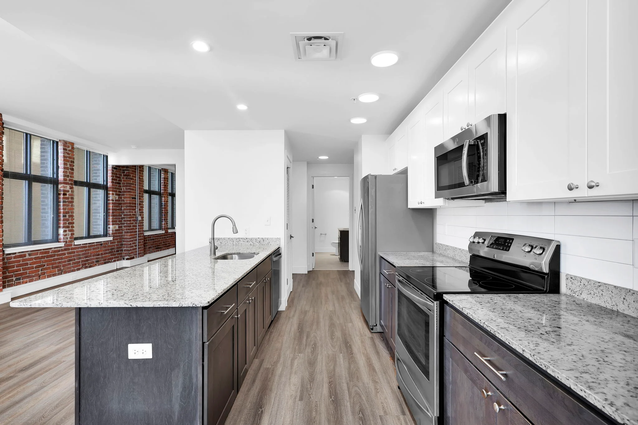 A modern kitchen featuring a large island with a granite countertop, dark wood cabinetry, stainless steel appliances, and large windows providing natural light.