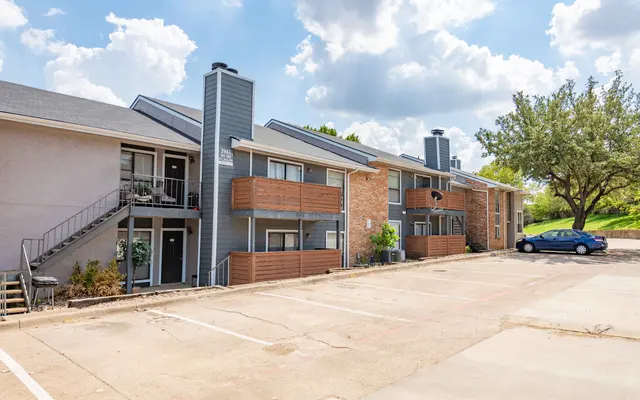 Exterior view of an apartment complex featuring two buildings, balconies, and a parking area with a blue car.
