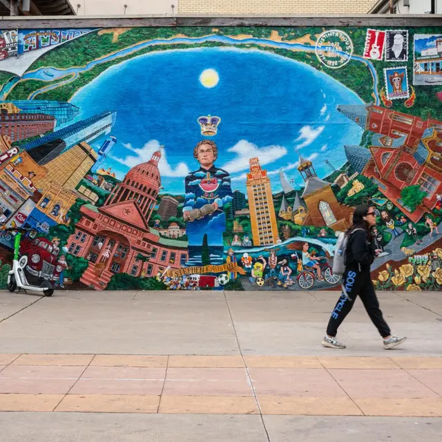 A colorful mural on a brick wall depicting a vibrant scene with buildings, characters, and abstract designs under a blue sky with a sun.
