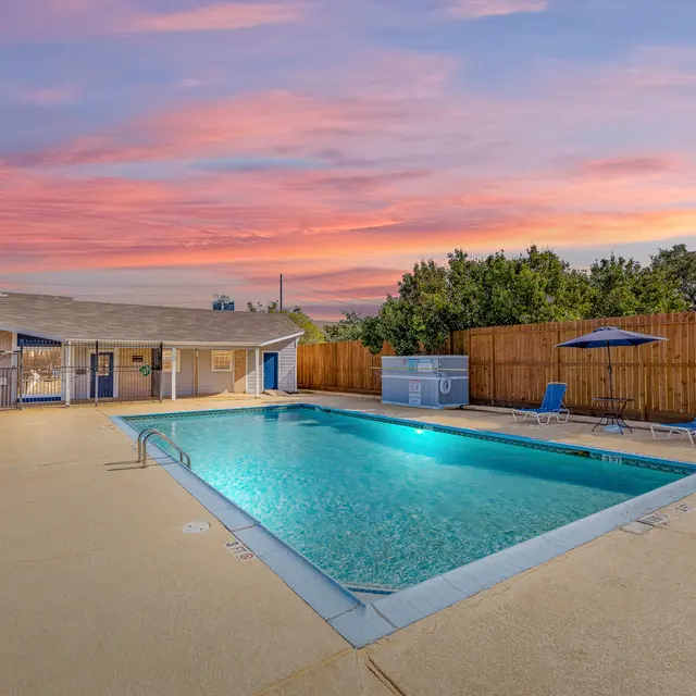 Swimming pool surrounded by a fence, with chairs and umbrellas, under a pink and blue sunset sky.