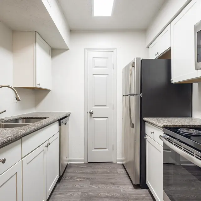 A modern kitchen featuring white cabinets, granite countertops, and stainless steel appliances. The kitchen includes a sink, stove, refrigerator, and a door leading to another room.