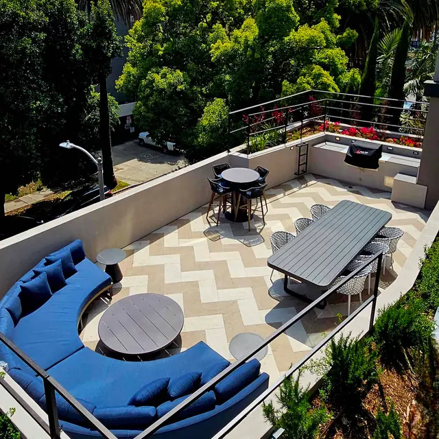 Chic Rooftop Lounge Area A spacious rooftop lounge area featuring a blue sectional sofa, a round coffee table, a long dining table, and bar stools, surrounded by greenery and large palm trees.