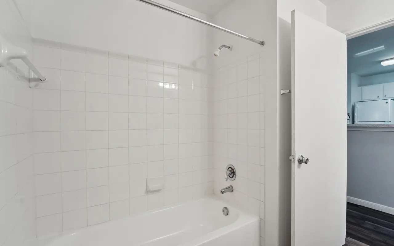 A bathroom featuring a white tiled bathtub with a shower rod, a white wall in the background, and an open door leading to another room.