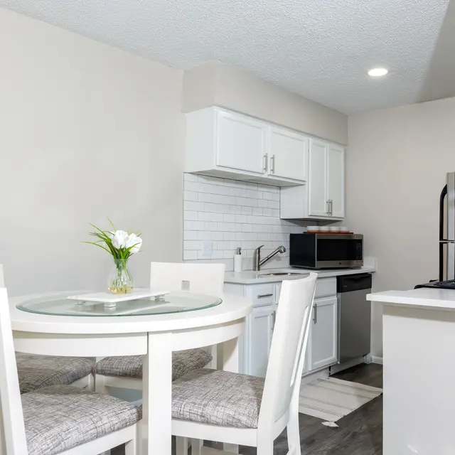 A modern kitchen with a dining table and chairs. The kitchen features white cabinetry, a microwave, and appliances. A glass-top dining table sits at the center, adorned with a small vase of flowers.