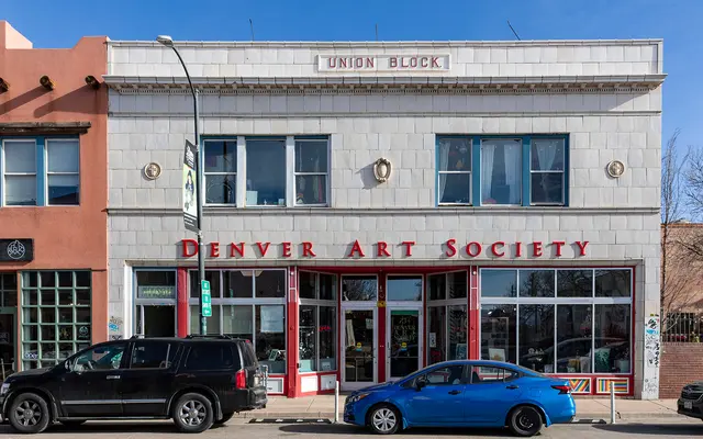 DENVER ART SOCIETY A two-story building with a sign that reads 'Denver Art Society'. The facade features large windows and decorative elements above. Two parked cars are visible in front, one black and one blue. In the background, there is a building with a sign that partially reads 'UNION BLOCK'.