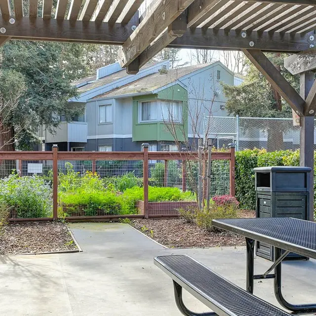 View of a shaded pavilion area with a picnic table and a trash can, surrounded by greenery and backed by residential buildings.