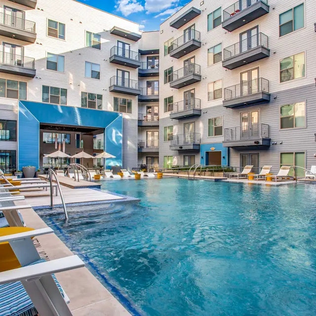 A luxurious apartment complex with a swimming pool in the center, surrounded by multiple floors of balconies. Lounge chairs are positioned near the pool, and blue skies are visible above.