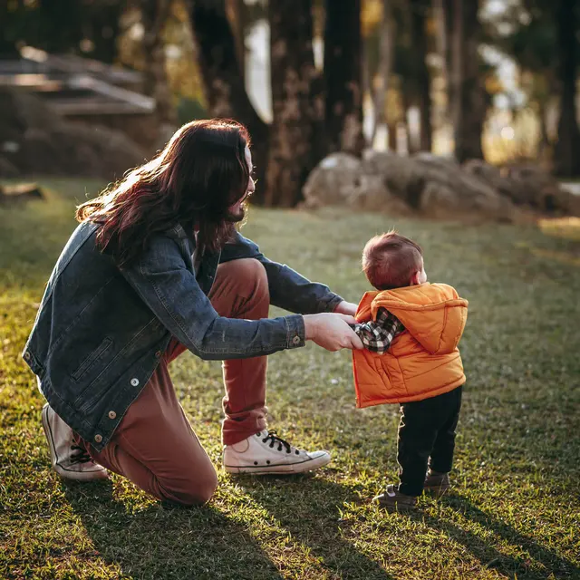 Mother and Child in Nature A woman kneeling on grass, reaching out to a small child who is standing and looking back. The child is wearing a vibrant orange jacket and black pants, while the woman is dressed in a denim jacket and light pants. The background features trees and rocks, creating a natural setting in soft sunlight.