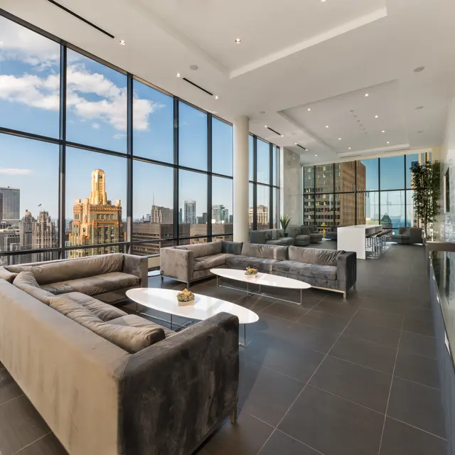 A modern loft-style living room with tall windows showcasing a city skyline. The room features gray sofas and a white coffee table on dark tiled flooring.