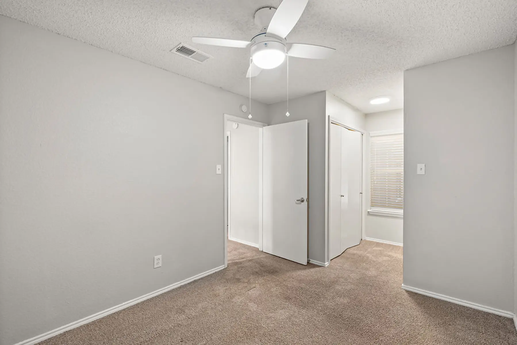 Minimalistic Bedroom Interior A clean and simple bedroom with beige carpet and light gray walls. The room features a light ceiling fan and two doors: one leading to a closet and another likely leading to another room. Natural light comes from a window on the opposite wall.