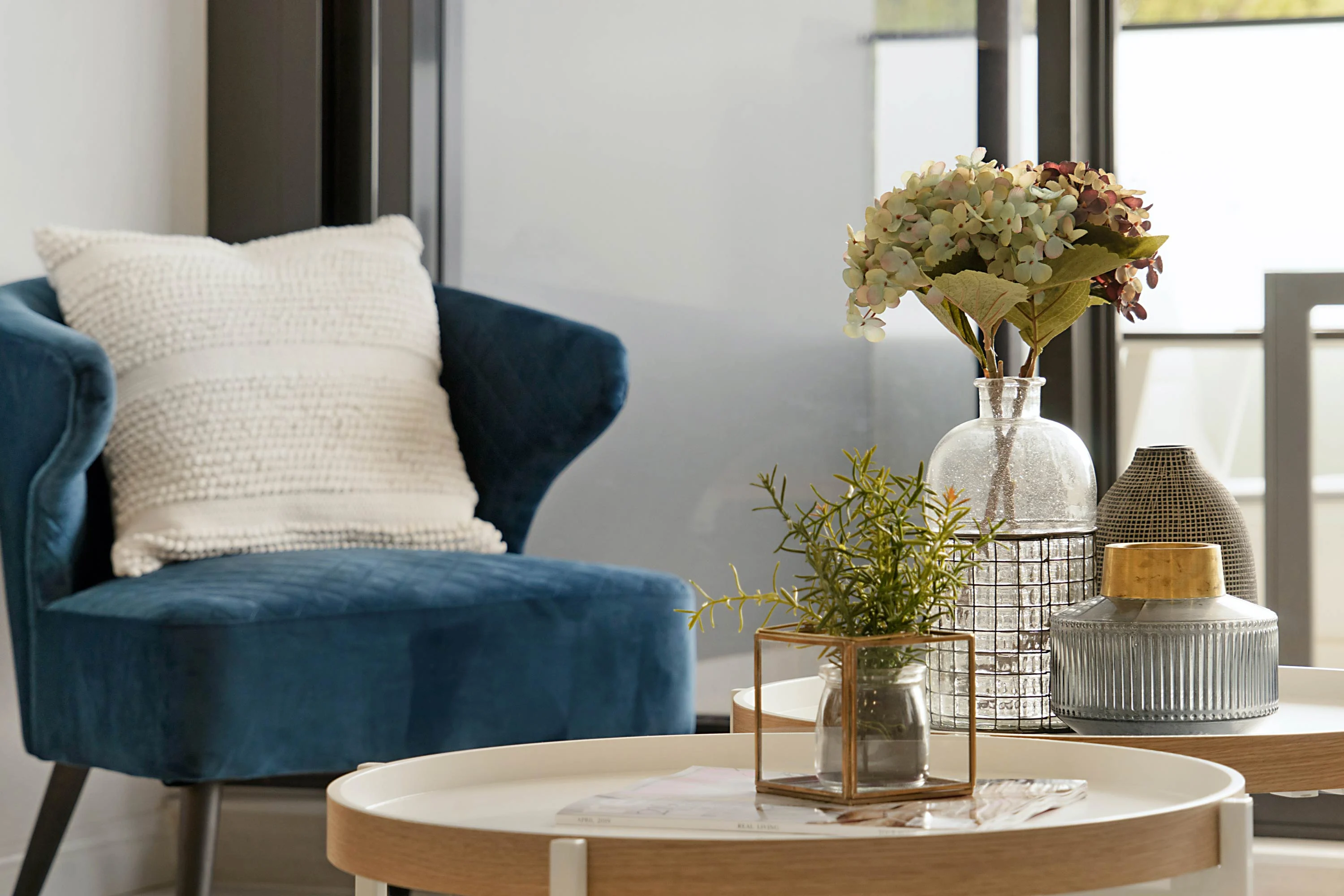 Cozy Living Room Decor A cozy living room scene featuring a blue armchair with a textured white pillow, a wooden coffee table with various decorative items including vases and a plant, and large windows allowing natural light to enter.