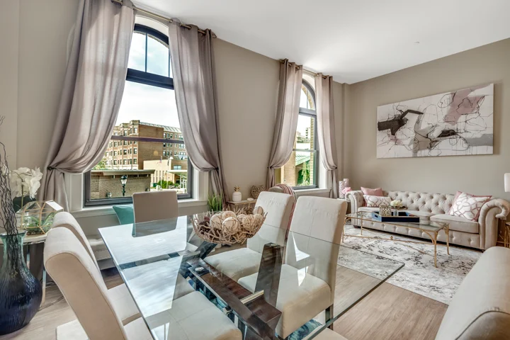 A stylish apartment interior featuring a glass dining table with beige chairs, plush sofas, and large windows with sheer curtains. Decor elements include a wall art piece and a soft area rug.