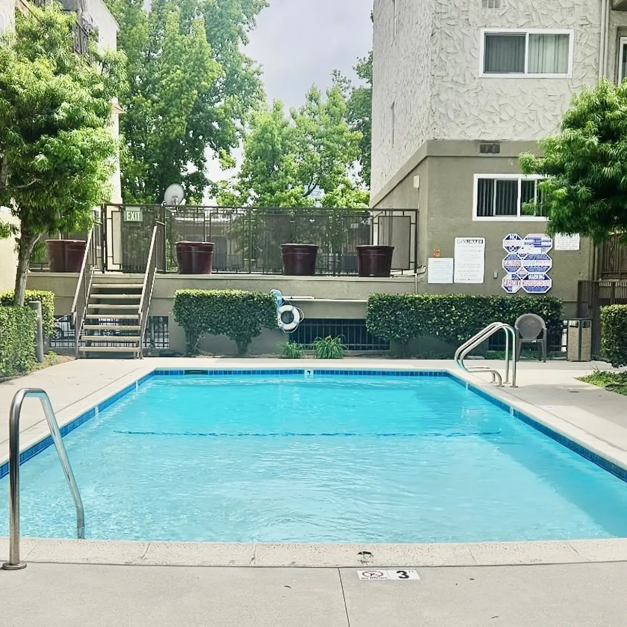 A clear swimming pool surrounded by greenery and apartment buildings under a cloudy sky.
