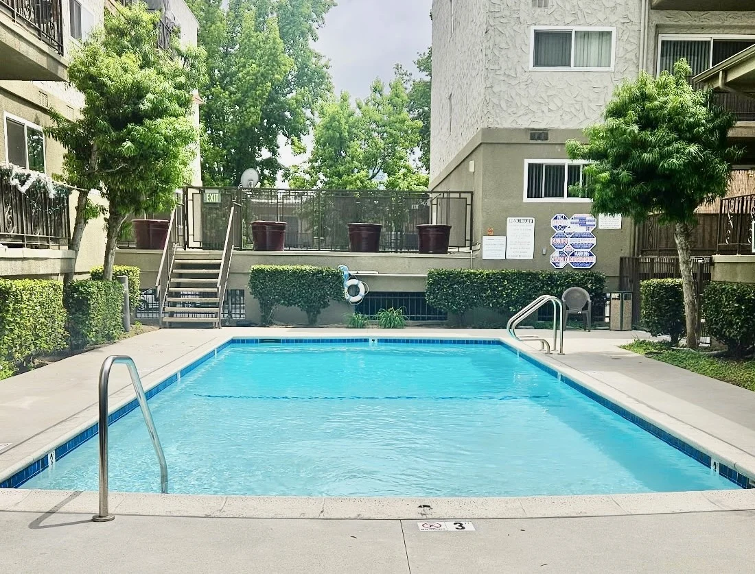 A clear swimming pool surrounded by greenery and apartment buildings under a cloudy sky.