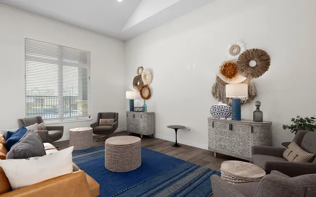 A stylish modern living room featuring two sets of comfortable armchairs, a brown leather sofa, and wooden coffee tables. The decor includes a blue area rug, a large window with blinds, and wall hangings made of natural materials. A decorative lamp and a small table add to the ambiance of the room, which has neutral-colored walls and abundant natural light.