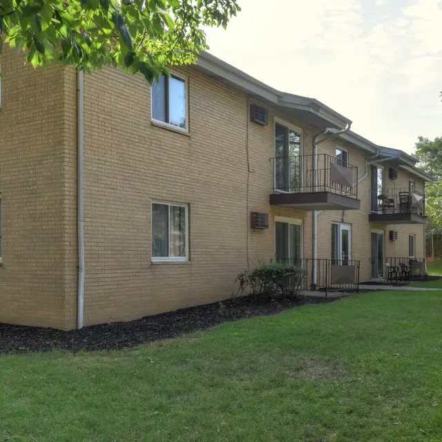 An exterior view of a multi-unit apartment building featuring a light-colored brick facade. The building has several windows and balconies, surrounded by a grassy area and trees.