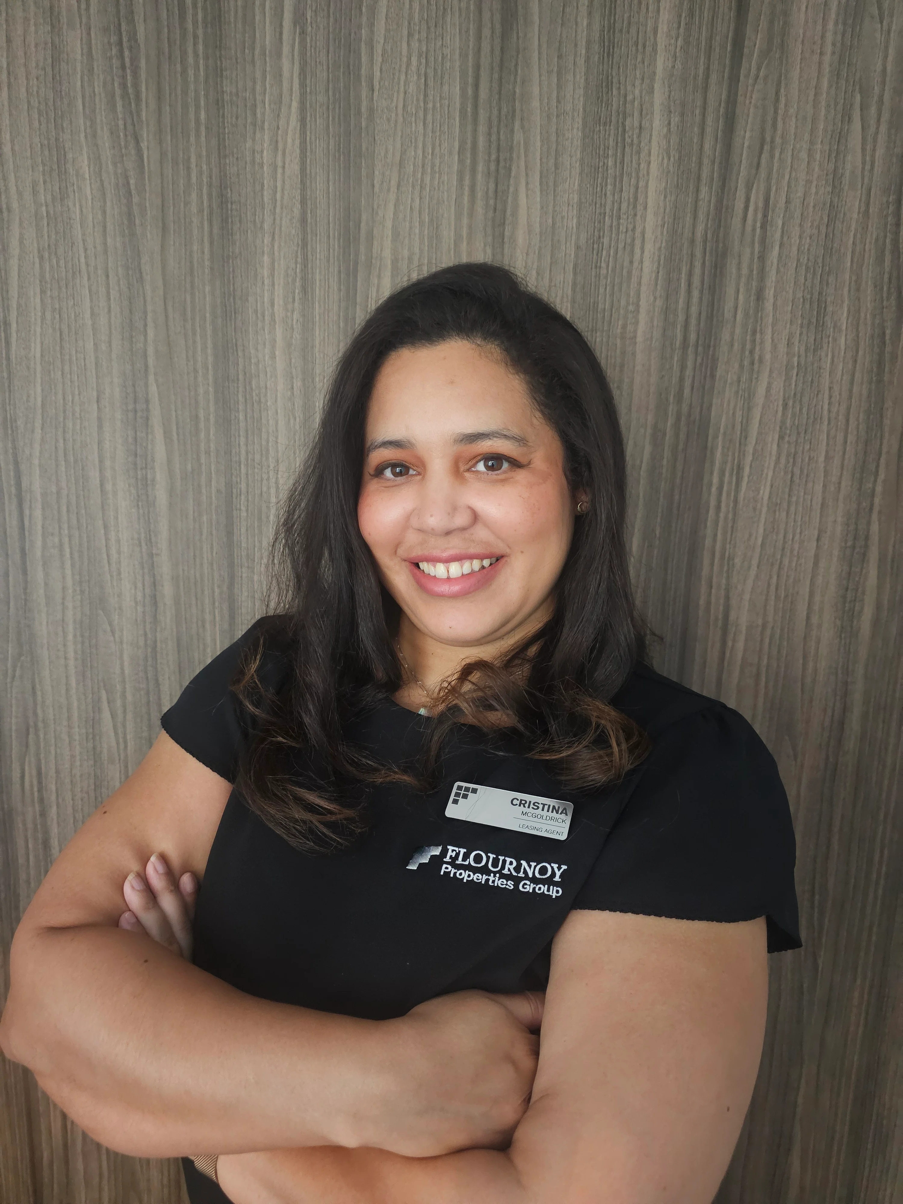 Professional Portrait of a Woman A smiling woman with long dark hair, wearing a black shirt, poses with her arms crossed in front of a wooden background.