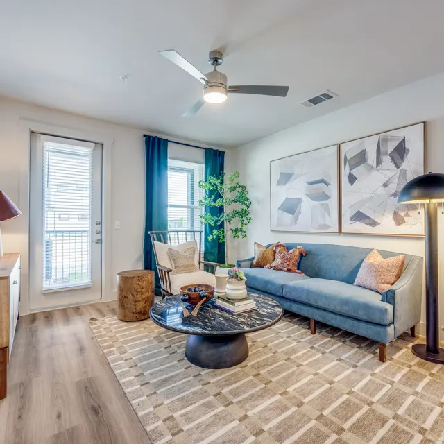 A modern living room with a blue sofa, decorative pillows, a large round coffee table, and artwork on the walls. A plant is in the corner and a lamp stands next to the sofa. There are wooden elements like a bookshelf and a side table, with a patterned rug on the floor.