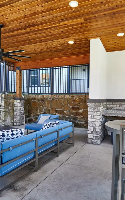 A modern outdoor living space featuring a wooden ceiling, stone walls, and comfortable blue furniture. There are two televisions mounted on the walls, and a small dining table with chairs. The area is well-lit with ceiling fans and natural light.