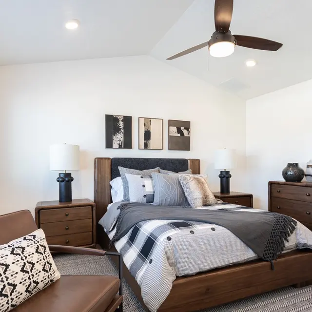 A modern bedroom featuring a double bed with decorative pillows, a ceiling fan, and a cozy armchair. The room includes wooden furniture and wall art.