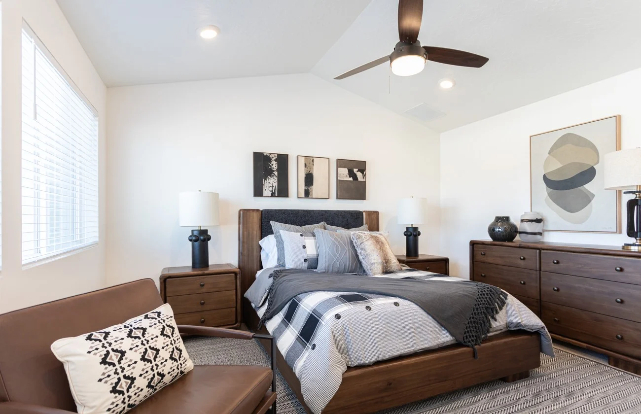 A modern bedroom featuring a double bed with decorative pillows, a ceiling fan, and a cozy armchair. The room includes wooden furniture and wall art.