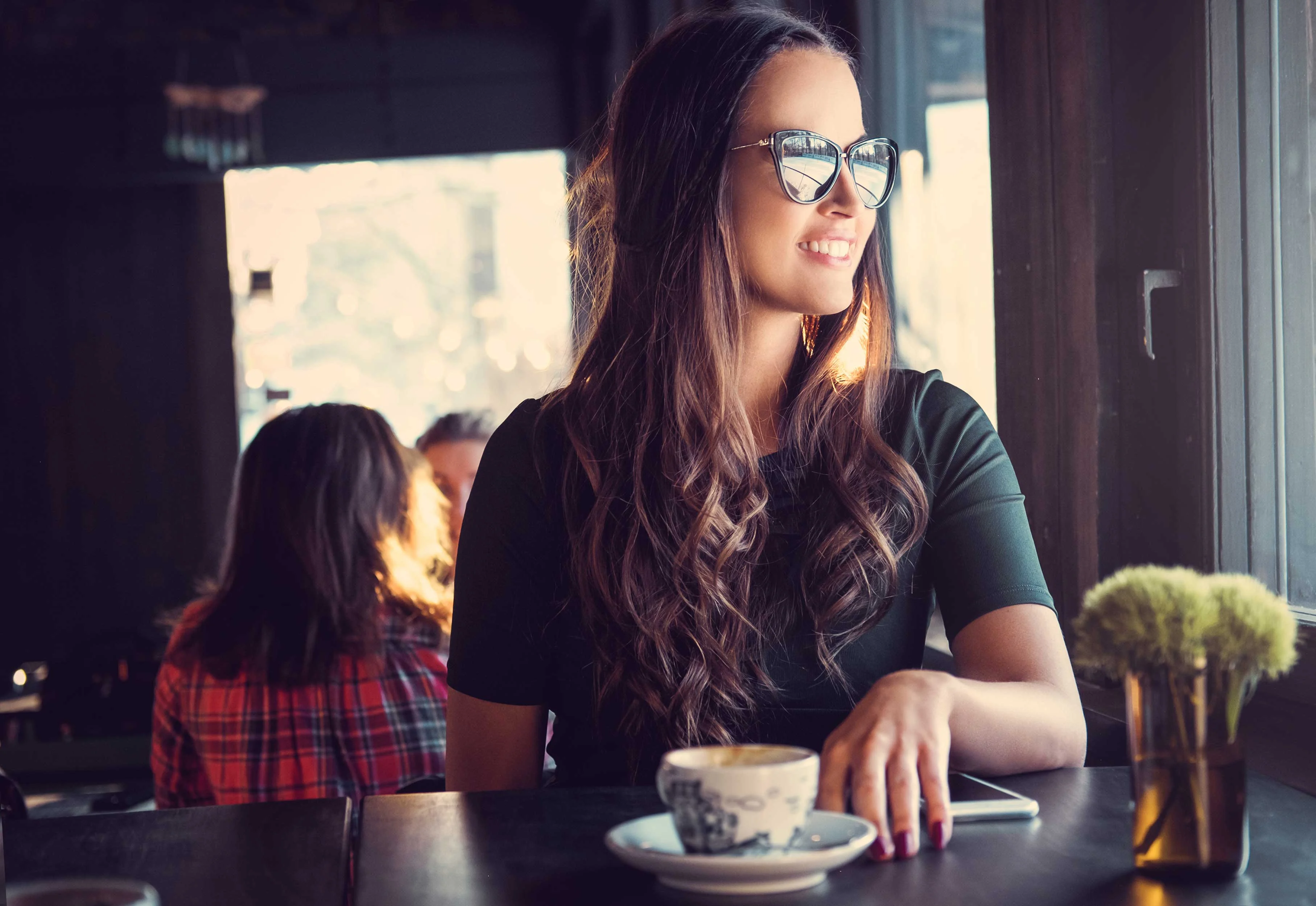 Charming Café Moment A woman with long hair and sunglasses sitting at a café table, gazing out the window. She has a cup on the table in front of her and looks content. In the background, another person is slightly visible, seated at a different table.