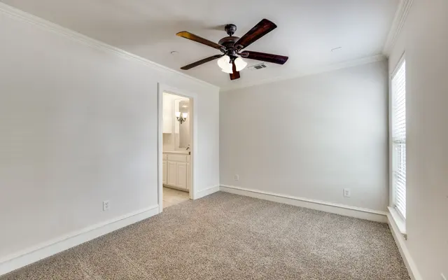 Spacious Bedroom Interior A spacious, well-lit bedroom featuring light-colored walls, a ceiling fan with wooden blades, and soft, plush carpet flooring. A doorway leads to an adjoining room, likely a bathroom, and there are windows on one wall with blinds.