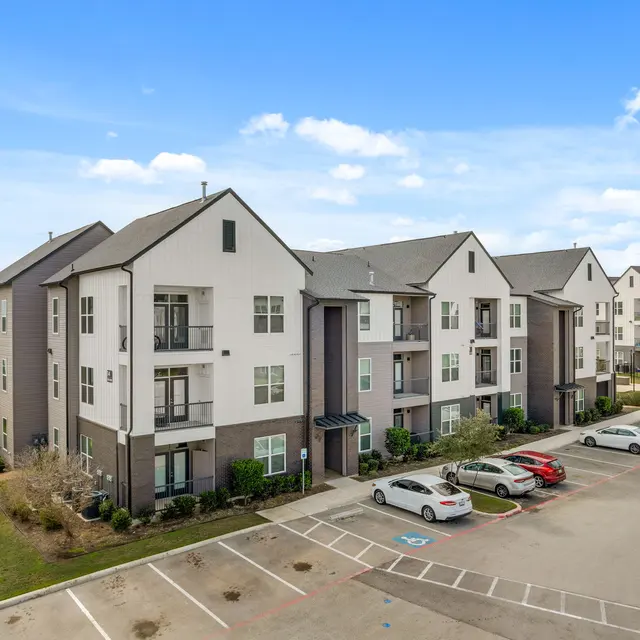 A modern apartment complex with multiple buildings, featuring a mix of light and dark colored exteriors. The parking lot is partially visible in the foreground with several cars parked.