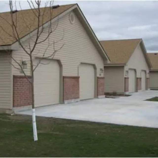 A row of beige residential houses with garage doors and small patches of grass in front. The sky is overcast, and a small tree is visible.