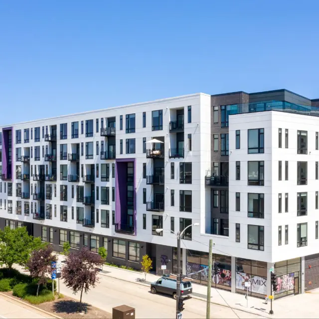 A modern multi-story apartment building featuring a blend of white and dark exterior with balconies, surrounded by trees and a sidewalk.